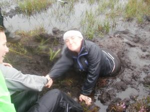 A person wearing outdoor clothing and a white beanie is stuck in muddy water, reaching up and holding hands with someone helping them out. Grassy and wet terrain surrounds them.