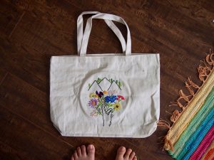 A white canvas tote bag with embroidered colorful flowers and mountains sits on a dark wooden floor near a rainbow-striped rug; bare toes are visible at the bottom of the image.