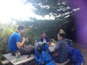 Three people sit at a wooden picnic table outdoors, smiling and talking. They have backpacks and a water bottle on the table. Trees and greenery surround them, suggesting a hiking or camping trip.
