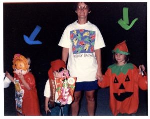 Three children in Halloween costumes stand with an adult. One child wears a Strawberry Shortcake mask, another is dressed as a pirate, and the third is in a pumpkin costume. The adult wears a Friday Harbor t-shirt.
