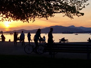 People relax near a waterfront at sunset, with silhouettes of walkers, cyclists, and sitters on benches. Mountains, boats, and a tree frame the scene against an orange sky.