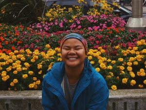A person in a blue jacket, smiling with eyes closed, sits in front of vibrant flower beds with yellow, orange, and pink flowers. A parked car and greenery are visible in the background.