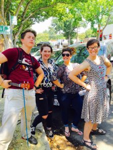 Four smiling people strike superhero poses on a sunny sidewalk near parked cars and trees; one person holds a small ukulele. All appear cheerful and playful.