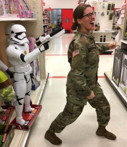 A woman in military uniform poses playfully as if running away while a life-sized Stormtrooper toy stands behind her, appearing to aim its blaster at her in a store aisle.