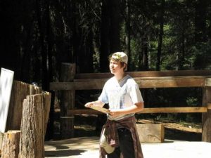A person wearing a cap stands outdoors in a sunlit wooded area, holding a clipboard. They have a shirt tied around their waist and are surrounded by wooden logs and a fence.