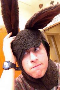 A person wearing a fuzzy brown costume with large floppy ears, resembling a rabbit or animal, looks upward while holding one ear upright. They have a watch on their wrist and are indoors.