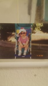 A young child sits in a stroller wearing a large white sunhat, pink shirt, pink sunglasses, and white socks with sneakers. The date 4-18-99 is visible in the lower right corner.
