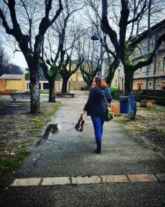 A woman walks away down a tree-lined path in a quiet courtyard, holding a violin and carrying a green bag. The ground is wet, and bare trees frame the scene on a cloudy day.