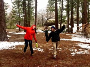 Two people in winter jackets and boots playfully pose in a snowy forest, arms outstretched and smiling. A third person stands in the background among tall pine trees and patches of snow.