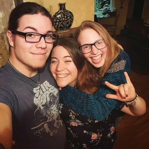 Three friends pose for a selfie indoors. The man on the left wears glasses and a Star Wars shirt. The two women, also wearing glasses, smile; one hugs the other from behind and makes a rock hand gesture. All look happy.