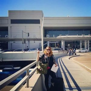 A woman stands outside an airport terminal, holding up one finger and smiling. She has luggage and a backpack beside her. The terminal building and several cars are visible in the background under a clear blue sky.