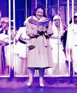 A woman in a tan coat stands in front of vertical bars, reading a book, with people dressed in white costumes behind her, on a stage with purple lighting.