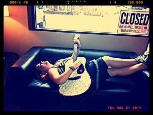 A young woman lies on a black couch playing a white guitar, smiling. She wears a striped top and black skirt. A “CLOSED” sign is visible in the window behind her. The photo has a retro filter and date stamp of August 21, 2014.