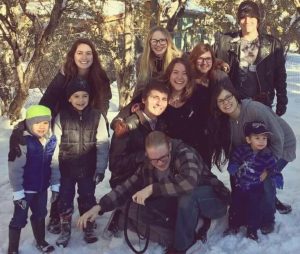 A group of adults and children pose together, smiling in a snowy outdoor setting with trees in the background. The children wear winter clothes and hats, and everyone looks happy.