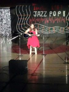 A woman in a red dress plays an acoustic guitar and sings on stage, sitting on a stool. Behind her is a black backdrop with colorful lines and the words JAZZ POP! in bold letters. Microphones are set up around her.