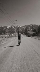 A person rides a bicycle down an empty, paved road toward distant snow-capped mountains, with houses, trees, and telephone poles lining the road under a clear sky.