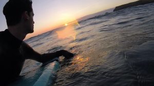 A surfer in a wetsuit sits on a surfboard in the ocean at sunset, looking toward the horizon as the sun sets over the water, creating reflections and a warm glow on the waves.