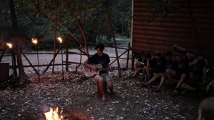A barefoot person plays guitar near a campfire outside a wooden cabin at dusk, while a group of people sit together nearby, surrounded by trees and rustic fencing.