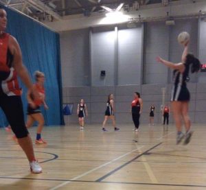 A group of women are playing an indoor netball game. One player in a black and white uniform is jumping to catch the ball, while others in red and black uniforms watch or move on the court. The court is lit with overhead lights.