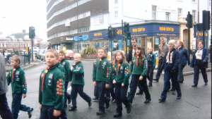 A group of children in green scout uniforms with badges walk in formation on a city street, accompanied by adults. Office buildings and shops are visible in the background.