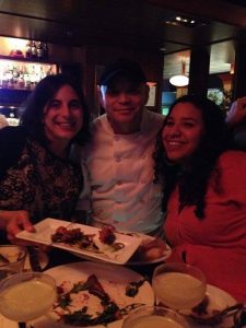 Two women smile at a table in a dimly lit restaurant, posing with a chef in a white uniform. Plates of food and drinks are in front of them, and the atmosphere appears warm and lively.