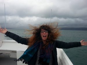 A woman with long, wind-blown hair and glasses smiles with arms outstretched on a boat, with cloudy skies and choppy water in the background.