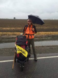 A person in an orange jacket stands on a wet road next to a stroller with a yellow Walking America sign, holding an umbrella in rainy weather, with farmland in the background.