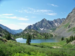 A clear mountain lake surrounded by pine trees and lush green grass, set beneath rugged, rocky peaks partially dotted with snow under a blue sky with scattered clouds.