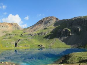 Clear blue lake reflects green hills and rocky mountain peaks under a bright blue sky with scattered clouds. The landscape appears serene and untouched, with sunlight illuminating the scene.