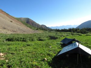 Two small tents are set up on a grassy meadow surrounded by green hills and distant mountains under a clear blue sky. Sunlight illuminates the landscape, suggesting early morning or late afternoon.