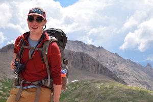 A smiling hiker wearing sunglasses, a cap, and a red jacket stands on a mountain trail. He carries a large backpack and holds a camera, with rocky peaks and cloudy sky in the background.