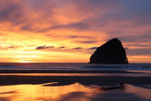A large, dark sea stack rises from the ocean near the shore at sunset, with golden and orange clouds reflected in wet sand and calm waves in the background.