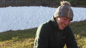 A person wearing a knit hat and green jacket smiles while sitting on grass, with a patch of snow and rocky ground in the background.