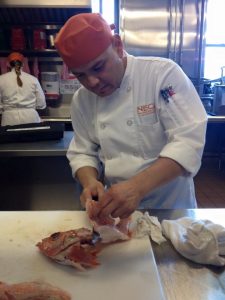 A chef in a white uniform and orange hat fillets a fish on a white cutting board in a professional kitchen. Another person in the background works at a counter.