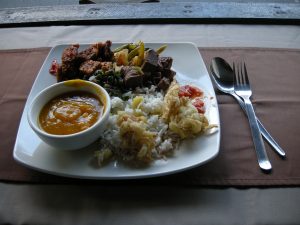 A square white plate with rice, fried vegetables, mixed meats, assorted vegetables, and a bowl of orange soup sits on a table with a fork and spoon on a napkin beside it.