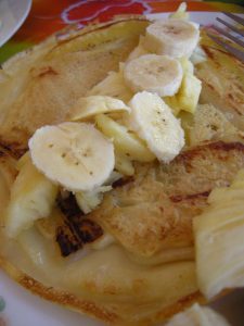 A close-up of a crepe topped with sliced bananas and pieces of pineapple on a plate, with a fork visible to the side.