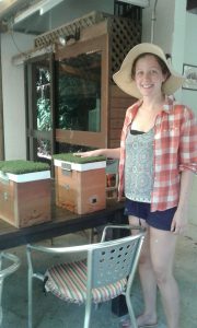 A woman in a sun hat and plaid shirt stands smiling next to two wooden boxes with green tops on a patio table, outdoors in a shaded area.