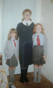 Three smiling children in school uniforms stand indoors. Two younger girls wear grey skirts, cardigans, red ties, and knee-high socks; the older girl wears a darker skirt and sweater with a tie and glasses. All have light brown or red hair.