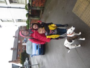 A woman in a red raincoat smiles with two children, one in a yellow jacket and the other hugging him, standing on a wet sidewalk with a white and brown dog on a leash. A blue car and brick buildings are in the background.