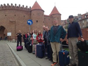 A group of ten people with suitcases stand on a sidewalk in front of a historic brick fortress with round towers and red roofs, near a blue left-turn traffic sign.