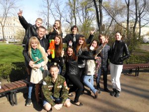 A group of thirteen young people pose outdoors in a park, many flashing peace signs and smiling. Some wear leather jackets, and one person holds colorful water guns. Trees and buildings are visible in the background.