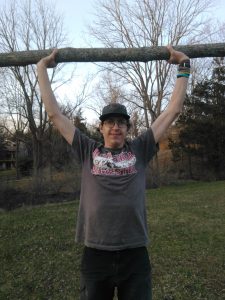 A person wearing glasses, a cap, and a t-shirt holds a large tree branch above their head with both hands, standing outdoors on grass with leafless trees in the background.