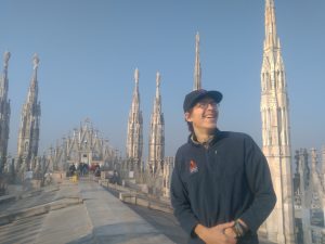 A smiling person in a dark jacket and cap stands on the rooftop of a cathedral with ornate spires, under a clear blue sky. Other people are visible in the background, admiring the architecture.