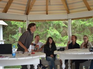 A group of five people sit and laugh together under a gazebo surrounded by trees. Four are seated at a white table, while one person stands, smiling and gesturing enthusiastically. Papers and a water bottle are on the table.