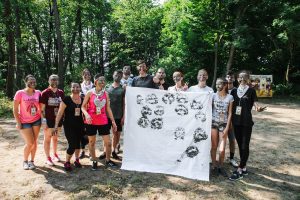 A group of people with painted faces stand outdoors on dirt, holding a large white sheet with black handprints. They are smiling, and trees fill the background on a sunny day.
