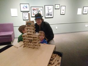 A man and two children sit behind a tall wooden block tower they built on a table, in a room with framed photos on the wall and a purple couch to the side.