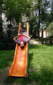 A man lies upside down on an orange playground slide in a park, with his legs up and head near the bottom, surrounded by green grass and trees.