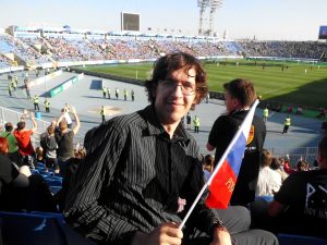 A man with glasses sits in a stadium seat, smiling and holding a small Russian flag. Behind him, there are many spectators, a soccer field, security personnel, and a clear sky.
