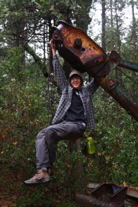 A smiling person in outdoor clothing hangs from a rusty piece of heavy machinery in a forest, holding on with both hands. They have a green water bottle attached to their pants and trees surround them.