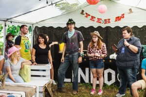 A group of people, some wearing cowboy hats, stand and sit in a decorated tent with YEEHAW banner overhead. One person holds a drink tray, while another reads from a phone. The atmosphere is festive and casual.